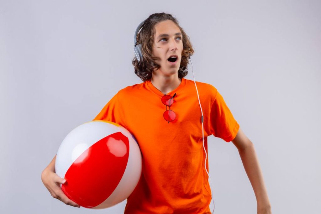 young-handsome-guy-orange-t-shirt-with-headphones-holding-inflatable-ball-looking-aside-surprised-amazed-standing